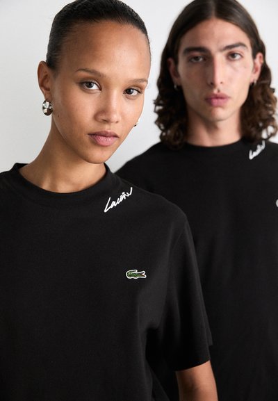 Крупен план на жена и мъж, носещи черни тениски Lacoste с логото на зелен крокодил и подписа близо до яката.