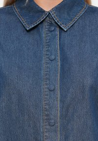 Jeansskjorte med tradisjonell krage, fem knapper og kontraststing. Stoffer er myke med en middels blå farge.