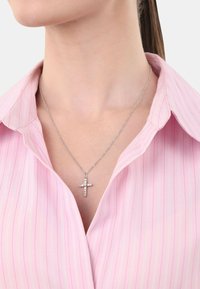 Collar de plata con un colgante de cruz adornado con pequeñas piedras transparentes, llevado sobre una camisa con cuello a rayas en color rosa claro.