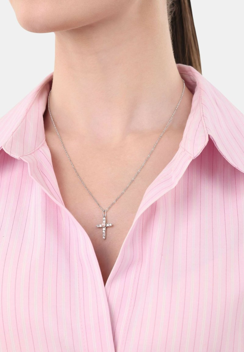 Collar de plata con un colgante de cruz adornado con pequeñas piedras transparentes, llevado sobre una camisa con cuello a rayas en color rosa claro.