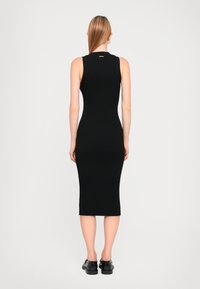 Robe midi noire côtelée avec un design sans manches, silhouette ajustée, col rond et un détail de marque remarquable au dos. Chaussures noires portées.