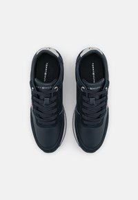 Ett par marinblå Tommy Hilfiger-sneakers med snören, vita mellansulor och varumärkesinsoler, vy ovanifrån mot en vit bakgrund.