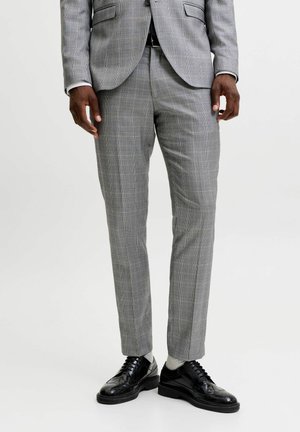 Homme portant un pantalon et une veste de costume à carreaux gris clair avec des chaussures habillées noires et des chaussettes claires, debout devant un fond uni.
