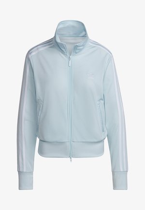 Lichtblauw Adidas trainingspak met witte strepen op de mouwen en broek, zakken met ritsen, en een persoon met handen in de zakken.
