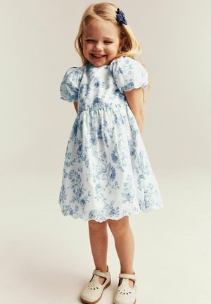 Lachend jong meisje in een witte jurk met blauwe bloemenpatronen, beige schoenen en een blauwe haarclip, staand met haar handen achter haar rug.