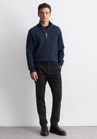 Marineblauer Strickpullover mit einem halben Reißverschluss-Kragen, kombiniert mit schwarzen Hosen und schwarzen Schuhen. Der Pullover hat gerippte Bündchen und Säume.