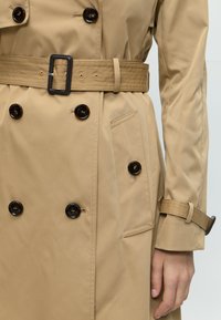 Beige trench coat with double-breasted dark buttons, matching belt with rectangular buckle, buttoned pocket, and cuff strap on right arm.