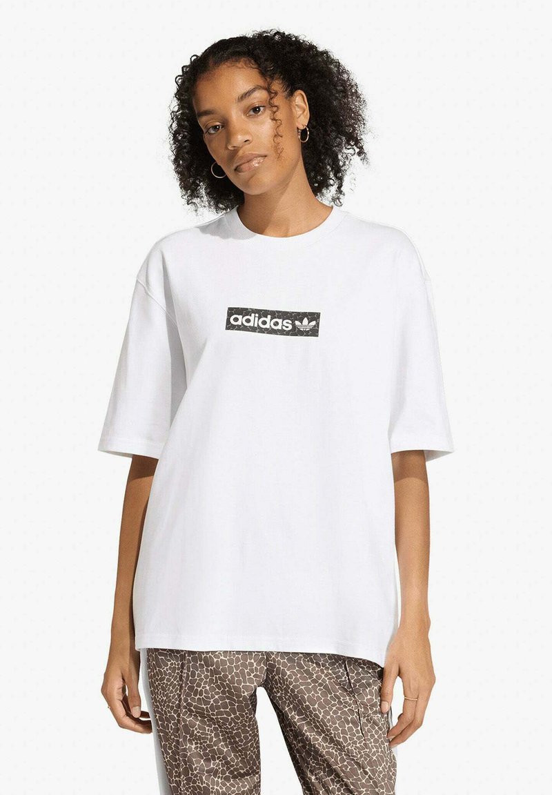 Giovane donna con capelli ricci che indossa una maglietta bianca Adidas e pantaloni marroni a motivi, in piedi davanti a uno sfondo chiaro e uniforme.