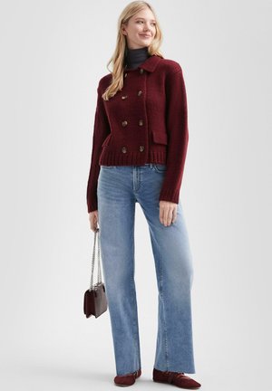 Cardigan en tricot bourgogne avec des boutons croisés, doté de deux poches et d'un ourlet côtelé, associé à un jean flare bleu clair et des chaussures bourgogne.