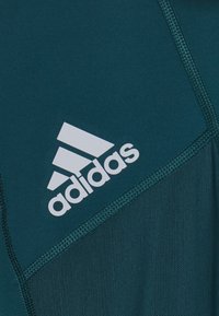 Teallinen urheilupaita, jossa on sileä pinta, verkko-osioita ja suuri valkoinen Adidas-logo vasemmalla puolella.