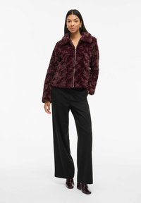 Giacca in pelliccia ecologica bordeaux con colletto e chiusura frontale con zip, abbinata a pantaloni neri a gamba larga e stivali con tacco bordeaux.