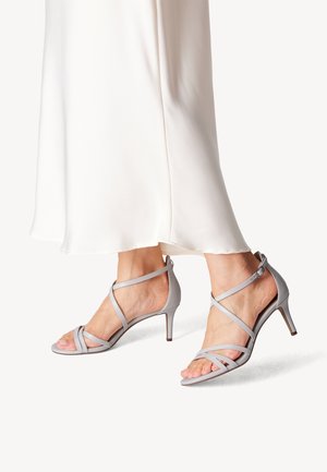 Szare sandały na wysokim obcasie z odkrytymi palcami, z wieloma skórzanymi paskami i smukłym, skośnym obcasem. Noszone z białą sukienką.