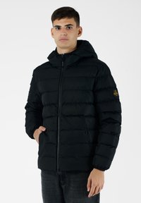 Giacca nera imbottita con chiusura a zip e cappuccio. Include polsini elastici, due tasche laterali e una toppa con logo sul braccio sinistro.