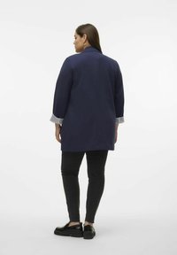 Navyblaues, langes Jackett mit Dreiviertelärmeln und gestreiftem Innenbund. Kombiniert mit schwarzen Slim-Fit-Hosen und Plateauschuhen.