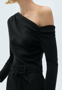 Schulterfreies, schwarzes Strickoberteil mit langen Ärmeln, strukturierter Oberfläche und gerafftem Detail am Ausschnitt. Kombiniert mit schwarzen Hosen.