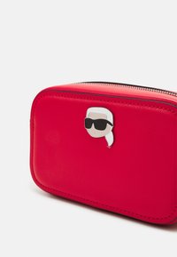 Czerwona skórzana prostokątna torba o gładkiej teksturze, z metalowym logo w stylu kreskówkowym z przodu i kontrastowym czarnym zamkiem błyskawicznym.