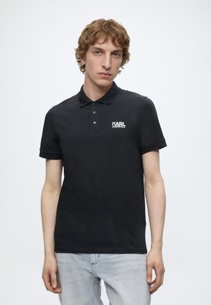 Junger Mann mit lockigem Haar, der ein schwarzes Karl Lagerfeld Poloshirt und hellgraue Jeans vor einem einfarbigen weißen Hintergrund trägt.