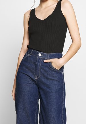 Femme portant un haut sans manches noir côtelé et un jean taille haute bleu foncé avec des coutures visibles, la main dans la poche.