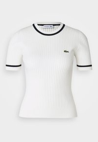 Bílý žebrovaný top s krátkým rukávem s námořnicky modrým lemováním na límci a rukávech. Na prsou je malé zelené logo krokodýla Lacoste.
