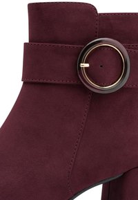 Stivaletti in suede bordeaux con risvolto ampio, cuciture dettagliate e una fibbia rotonda con accento dorato per la chiusura. Texture liscia in tutto il prodotto.