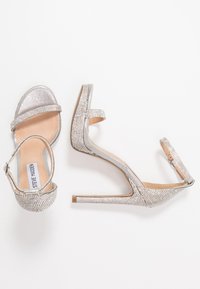 Stříbrné vysoké sandály na podpatku s designem stiletto, s třpytivým páskem zdobeným kamínky a hladkým metalickým povrchem.