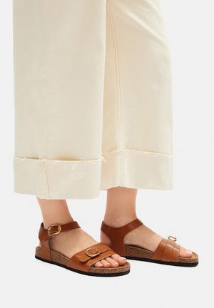 Brązowe skórzane sandały z klamrą i paskiem, wyposażone w korkową wkładkę i czarną gumową podeszwę. Noszone z jasnobeżowymi, krótkimi spodniami.