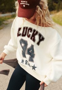 Biely oversized sveter s nápisom "LUCKY 94" v tmavom písmene, so vzormi hviezd; v kombinácii s čiernymi priliehavými legínami a hnedou čiapkou s nápisom.