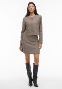 Žena stojící ve hnědém kostkovaném saku a odpovídající sukni s černými vysokými kozačkami na bílém pozadí.