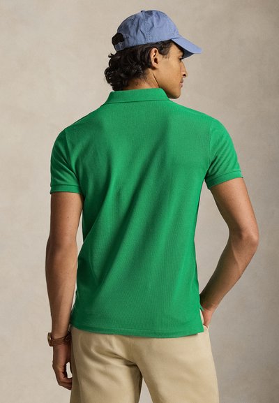 Άνδρας που φοράει πράσινο πόλο με κοντά μανίκια, μπεζ παντελόνι, μπλε καπέλο και καφέ ρολόι, στέκεται με την πλάτη προς την κάμερα μπροστά σε μπεζ φόντο.