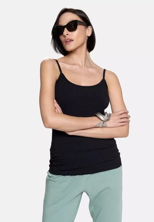 Czarny top na ramiączkach, zestawiony z jas green spodniami. Srebrna bransoleta na jednym ramieniu, modelka nosi ciemne okulary przeciwsłoneczne.