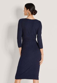 Frau mit dunklem Haar im Dutt, die ein enges, dunkelblaues, knielanges Kleid mit drei Viertelärmeln trägt und mit dem Rücken zur Kamera posiert.