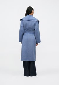 Hátulról látható nő, aki derékban megkötött kék kabátot visel széles gallérral és sötét, széles lábú nadrággal, egyszínű háttér előtt.