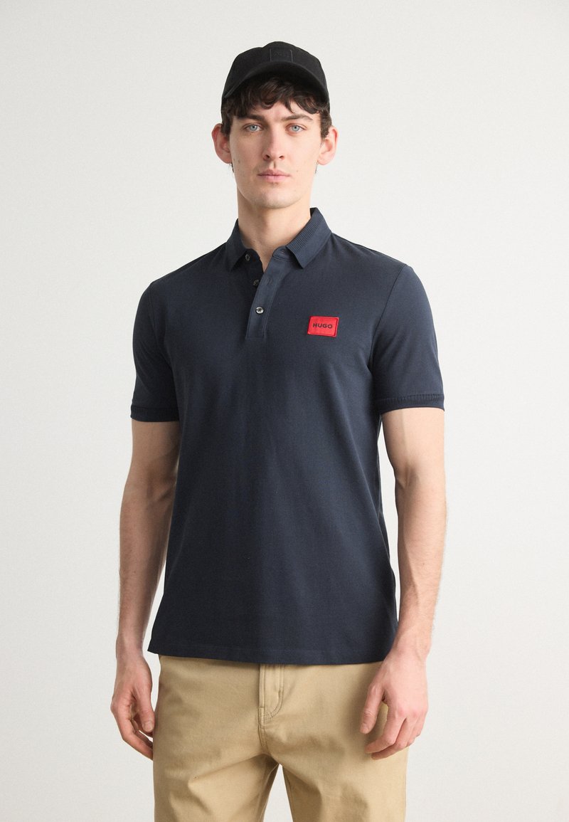 Marineblå polo med slim fit, korte ermer og en rød rektangulær logo-patch på brystet. Kombinert med beige bukser og en svart caps.