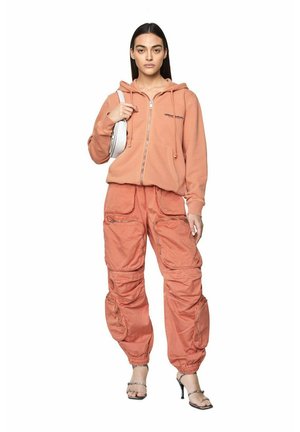 Donna che indossa una felpa arancione con cerniera e pantaloni cargo coordinati, tiene una borsa bianca a spalla e indossa sandali con tacco alto e punta aperta.