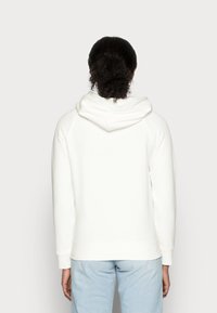 Weißer Hoodie mit großzügiger Kapuze, Raglanärmeln und gerippten Bündchen. Glatte Textur, schlichtes Design und ohne sichtbare Muster. Hellblaue Jeans.