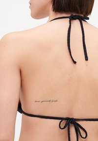 Svart halterneck-bikini topp med flettede stropper knyttet bak. En tatovering på øvre rygg viser teksten "elske deg selv først." Glatt hudtekstur.