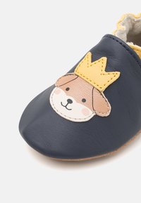 Chaussure pour bébé en cuir bleu marine avec une semelle texturée, présentant une appliqué de visage de chien avec une couronne jaune et des accents roses. Doublure intérieure douce.