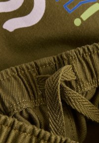 Ceinture élastique vert olive avec cordon plat. Le tissu doux et texturé présente un imprimé blanc et coloré sur le vêtement.