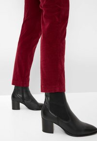 Schwarze Leder-Ankle-Boots mit Blockabsatz und elastischen Seitenpaneelen, kombiniert mit eng anliegenden burgunderfarbenen Cordhosen. Glattes Material und schnörkelloses Design.