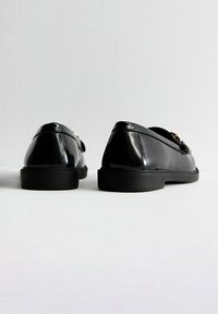 Mocassini neri in vernice con texture liscia, punta arrotondata, suola in gomma a profilo basso e dettaglio metallico sulla parte superiore.