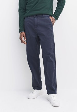 Mann trägt marineblaue gerade Hose, weiße Sneakers und ein grünes Langarmshirt mit gestreiften Bündchen, steht mit einer Hand in der Tasche.