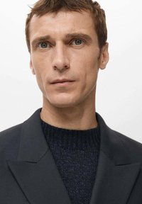 Modelo masculino con chaqueta oscura sobre un suéter azul marino texturizado, con cabello corto castaño y ojos azules, sobre un fondo claro.