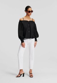 Blusa nera off-shoulder con maniche a sbuffo, chiusura con bottoni e dettagli in pizzo, abbinata a jeans bianchi a gamba dritta e sandali neri con tacco a blocco.