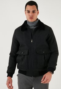Črna volnena bomberska jakna s črnim umetnim krznom okoli ovratnika, dvema sprednjima žepoma, rebrastimi manšetami in robom ter zadrgo. Gladka tekstura, minimalistična zasnova.