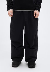 Μαύρο, φαρδύ παντελόνι με λεία υφή, με φαρδιά κοπή και ελαστική μέση, συνοδευόμενο από μαύρα παπούτσια.