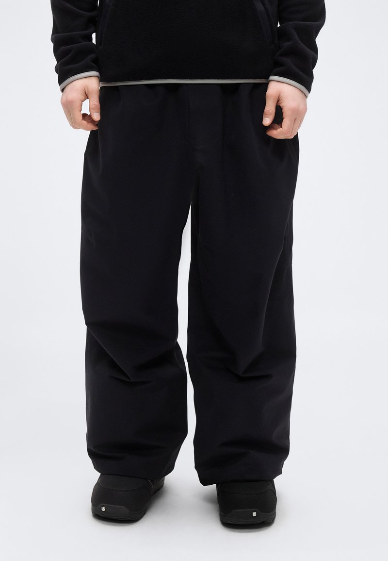 Μαύρο, φαρδύ παντελόνι με λεία υφή, με φαρδιά κοπή και ελαστική μέση, συνοδευόμενο από μαύρα παπούτσια.