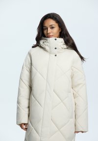 Ecrufarbener Puffer-Mantel mit einem Rautensteppmuster, hohem Kragen, versteckten Druckknöpfen und Seitentaschen. Glatte, glänzende Textur.