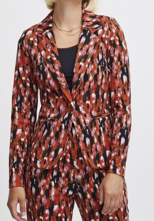 Mujer con una blazer de estampado en rojo, negro y blanco y pantalones a juego, con una camiseta negra debajo y un collar dorado.