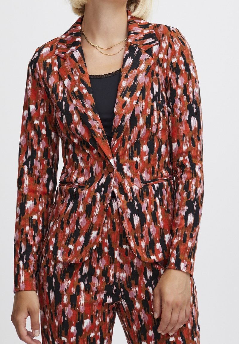 Femme portant un blazer à motifs rouges, noirs et blancs avec un pantalon assorti, un haut noir en dessous et un collier en or.