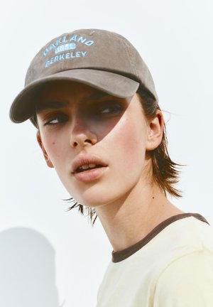 Młoda osoba z krótkimi włosami, nosząca beżową czapkę z niebieskim napisem „Oakland Berkeley” oraz jasną koszulę z ciemnym kołnierzem.